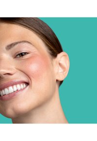 Gros plan d'une femme souriante à la peau lisse, maquillage léger, cheveux bruns tirés en arrière, sur un fond bleu sarcelle.