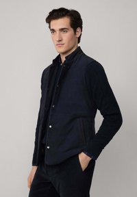 Tamnoplavi kaput s kvadratnom podlogom, prednje kopčanje na dugmad, dva bočna džepa, kontrastni detalji šavova; nosi se preko teksturirane, tamnoplave košulje s dugim rukavima.