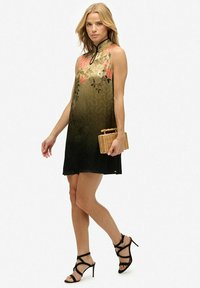 Femme portant une robe sans manches verte à fleurs ombrées, tenant une pochette en osier et des sandales à talons hauts à lanières noires, marchant devant un fond blanc.