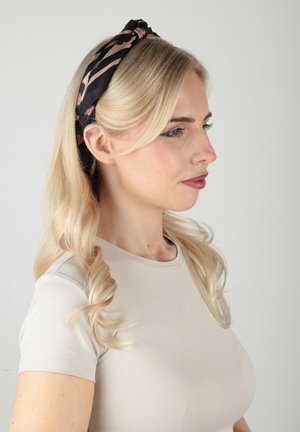 Donna bionda con capelli ricci indossa una fascia nera e marrone a fantasia e una maglietta beige a maniche corte, rivolta verso destra su uno sfondo grigio.