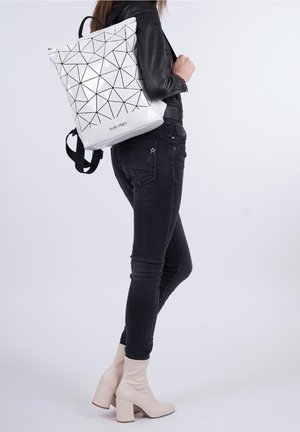 Personne en jean noir et veste portant un sac à dos SURI FREY blanc à motifs géométriques, chaussée de bottines beiges à talons.