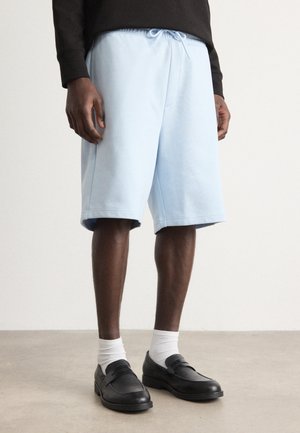 Lyseblå treningsshortser med snøring i livet, med en avslappet passform og sidesømmer, kombinert med svarte loafers og hvite sokker.