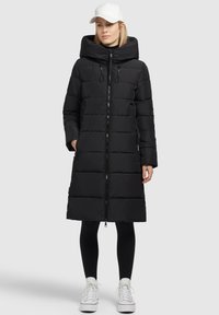 Zwarte gewatteerde jas met hoge kraag en ritssluiting aan de voorkant, met een capuchon en zijzakken, gedragen over zwarte leggings en witte sneakers.