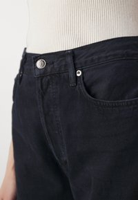 Nahaufnahme der Taille einer schwarzen Denim-Jeans mit Metallknopf und Niete, kombiniert mit einem weißen, gerippten Oberteil, das eingesteckt ist.
