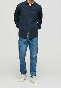 Camicia a maniche lunghe blu navy con una singola tasca sul petto, abbinata a jeans azzurri chiari e sneakers bianche. La camicia presenta una chiusura con bottoni.