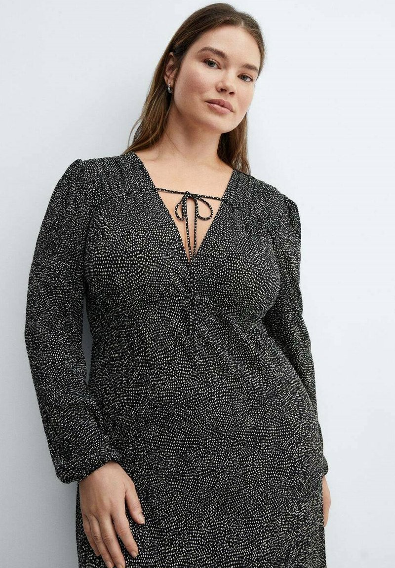 Giovane donna con capelli lunghi che indossa un vestito nero con un motivo a pois bianchi e un profondo scollo a V legato con un fiocco di stringa, in piedi contro uno sfondo chiaro.