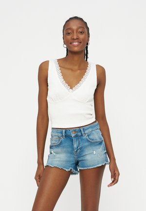 Νεαρή γυναίκα που χαμογελά, φορά λευκό αμάνικο τοπ με δαντέλα και φθαρμένο μπλε τζιν σορτς, σε απλή λευκή φόντο.
