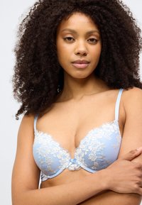 Lyseblå blonde-bh med blomsterbroderi, justerbare stropper og en lille central sløjfe. Tekstureret stof fremhæver visuelle detaljer.