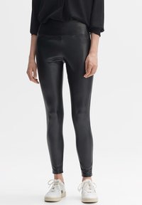 Svarta leggings i konstläder med hög midja, slät textur och figurnära design, kombinerat med vita sneakers för kontrast.