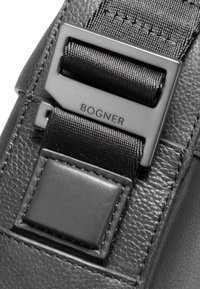 Detail černé kožené tašky s hladkou texturou, kovovým zapínáním s vyrytým nápisem "BOGNER" a odolným látkovým popruhem.
