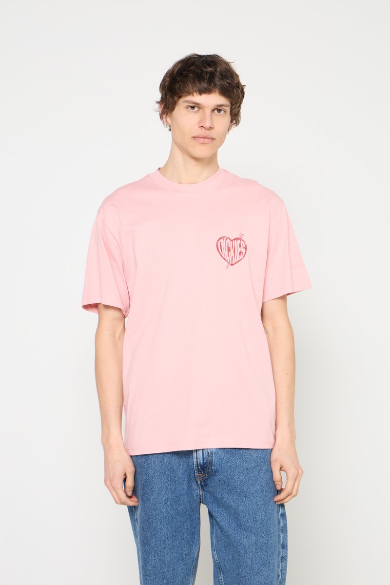 Jeune homme portant un T-shirt rose clair avec un cœur rouge et un texte sur la poitrine, associé à un jean bleu, debout devant un fond blanc uni.