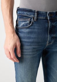 Jeanși din denim de culoare albastru închis, cu un finisaj ușor decolorat, cinci buzunare și elemente de bronz. Cusături de contrast vizibile de-a lungul cusăturilor și buzunarelor.