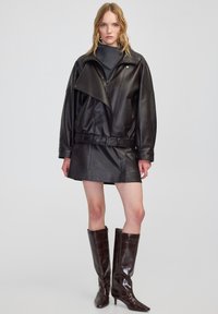 Giacca di pelle nera con vita cinturata, abbinata a una mini gonna di pelle nera. Stivali in pelle lucida marrone al ginocchio con punta affusolata.