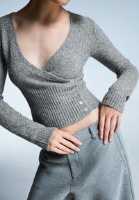 Szürke kötött kardigán V-alakú nyakkal, bordázott felülettel és gombos záródással. Világosszürke nadrággal párosítva. Puha, kényelmes anyag.