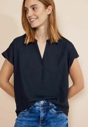 Jeune femme aux cheveux châtain clair mi-longs, portant un haut noir ample à col en V et manches courtes, ainsi qu'un jean bleu, souriante et regardant vers la gauche.