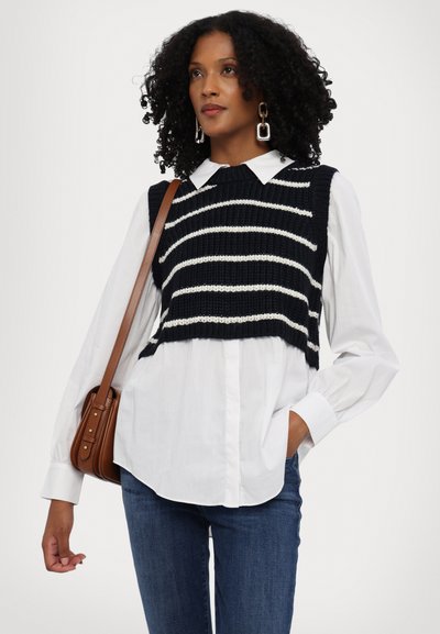 Navy gestricktes Crop Top mit weißen Streifen über einem weißen Hemd. Kombiniert mit blauen Jeans und einer braunen Umhängetasche mit goldenen Details.