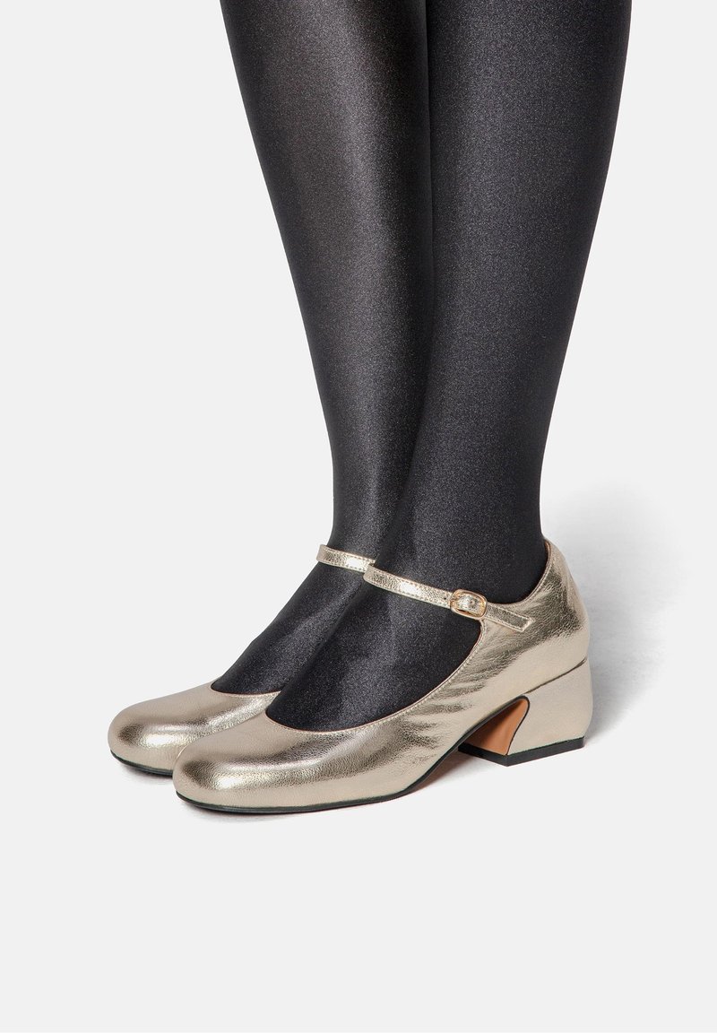 Chaussures Mary Jane métalliques dorées avec un bout arrondi, un petit talon carré et une bride décorative à la cheville. Portées avec des bas noirs brillants.