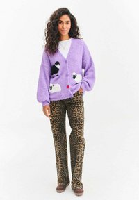 Kobieta z długimi kręconymi włosami, ubrana w fioletowy sweter z motywem owiec, spodnie w panterkę i brązowe buty, stojąca na białym tle.