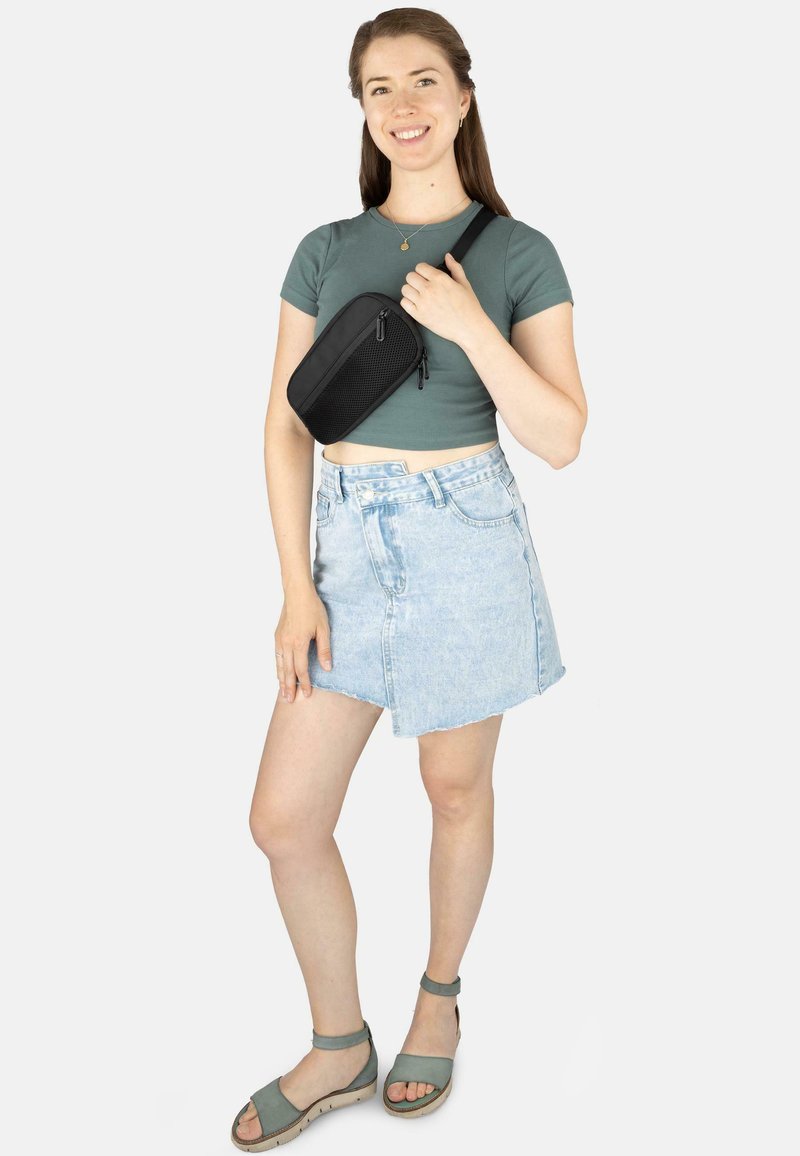 Schwarze, strukturierte Mini-Tasche, die mit einem Riemen gehalten wird, kombiniert mit einem engen grünen Oberteil und einem hellen Jeansrock, getragen mit grünen Sandalen.