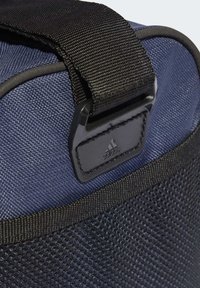 Borsa di stoffa blu con superficie testurizzata, tracolla nera regolabile e un'etichetta con logo nero prominente che mostra un design a tre strisce.