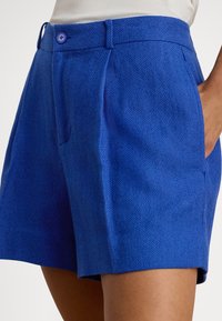 Blå fiskebensmønstrete shorts med glatt tekstur, som har en frontlomme og knappeåpning ved livlinningen.