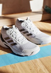 Białe buty sportowe z teksturowaną cholewką, trzema czarnymi paskami i wzmocnionym czubkiem. Posiadają amortyzowaną podeszwę dla wsparcia i przyczepności.