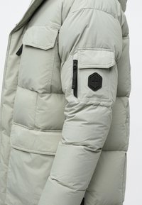 Ανοιχτό γκρι padded μπουφάν με κουκούλα. Διαθέτει τσέπη στο στήθος, τσέπη με φερμουάρ στον βραχίονα και εξαγωνική ετικέτα στον βραχίονα. Λείος υφασμάτινος χαρακτήρας.