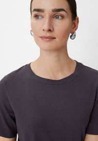 Czarnoszary t-shirt z okrągłym dekoltem, krótkie rękawy, gładka tekstura. Noszony z błyszczącymi, srebrnymi kolczykami w kształcie kropli, miękkie oświetlenie podkreślające rysy twarzy.