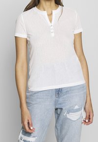 White short-sleeve henley shirt with a textured finish and three button placket. Paired with distressed light blue jeans.