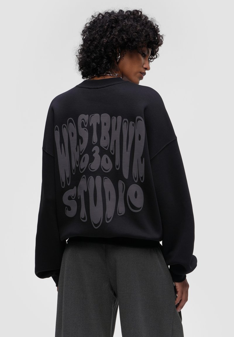 Felpa nera oversize realizzata in tessuto morbido, caratterizzata da una grande grafica stilizzata grigia sulla schiena con il testo "WRSTBHVR 030 STUDIO".