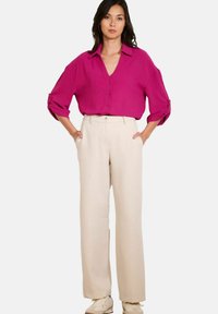 Vrouw in een magenta blouse met knopen en opgerolde mouwen, ingestopt in hoge beige wijde broek en beige sneakers.