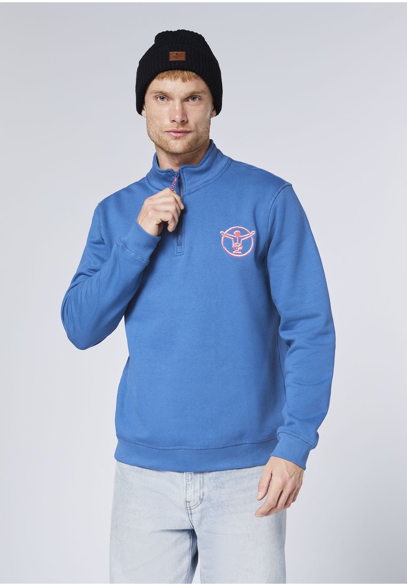 Blauer Fleecepullover mit halbem Reißverschluss am Kragen, orangefarbenem Logodesign auf der Brust, gerippten Bündchen und unterem Saum, getragen mit einer schwarzen Strickmütze.
