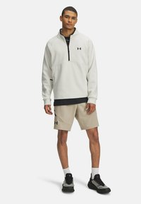 Gaišas krāsas vilnas džemperis ar ceturtdaļas rāvējslēdzēju, apvienots ar beidža šortiem. Iespējas melnie akcenti un Under Armour logotips. Sportisks piegriezums.