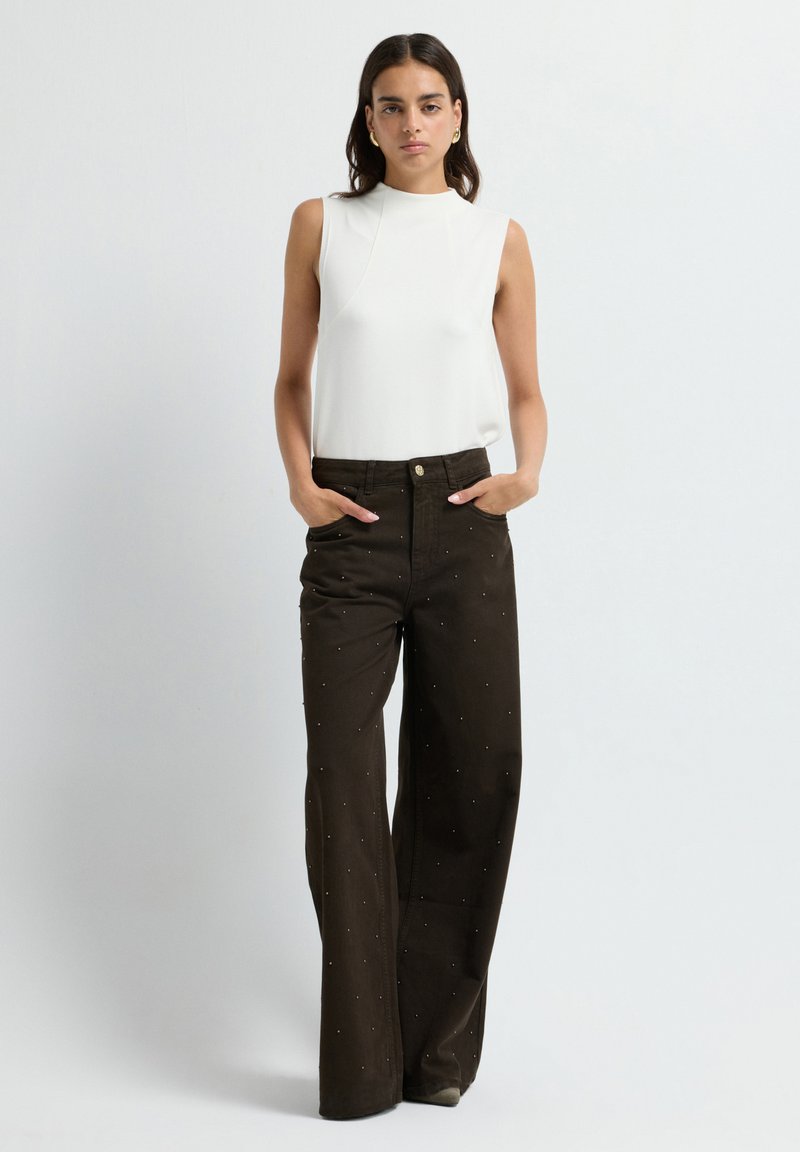 Haut sans manches blanc avec un col montant, accompagné de pantalons larges marron ornés de clous en argent et de poches avant.
