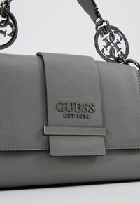 Sac à main gris Guess avec fermeture à rabat, détail barre métallique et breloques circulaires en chaîne fixées à la bandoulière.