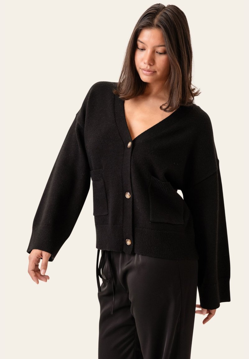 Mujer con cárdigan negro suelto con botones y bolsillos delanteros y pantalones negros, mirando hacia abajo con una postura relajada sobre un fondo liso.