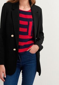 Nainen, jolla on päällään musta bleiseri, jossa on kultaiset napit, punavalkoraidallisen paidan ja sinisten farkkujen päällä, käsi taskussa, yksinkertaista taustaa vasten.