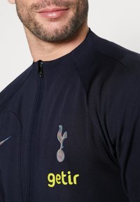 Giacca zip-up blu navy con texture liscia, dotata di un logo della squadra iridescente e testo giallo "getir" sulla parte anteriore.
