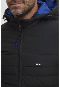 Svart puffjakke med blå hette, med glidelåslukking og horisontal sømdesign. Merkbar logo med røde, hvite og blå accenter.