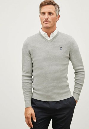 Grijze gebreide trui met een V-halsontwerp, geribde manchetten en zoom. Bevat een klein blauw embleem op de linkerborst. Gecombineerd met donkere broeken.