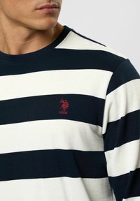 Close-up van een man die een marineblauw-wit gestreept shirt met lange mouwen draagt, met een rood USPA-logo geborduurd op de borst.