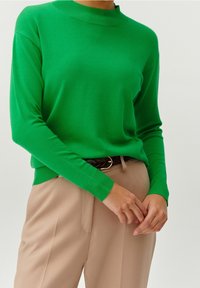 Personne portant un pull vert vif à manches longues rentré dans un pantalon beige avec une ceinture tressée foncée, se tenant devant un fond uni.