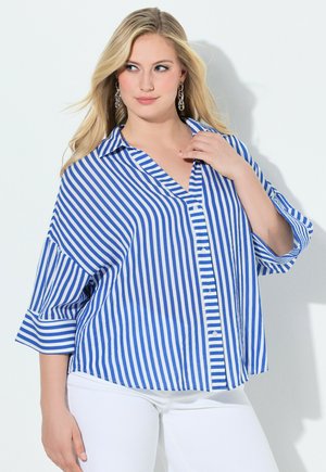 Frau trägt ein blau-weiß vertikal gestreiftes Hemd mit Knöpfen und 3/4-Ärmeln sowie weiße Hose, posiert vor einem schlichten hellen Hintergrund.