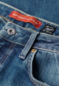 Vaqueros de mezclilla en azul con costuras en amarillo contrastante, presentando un botón plateado y etiquetas de marca en rojo y negro para algodón orgánico.