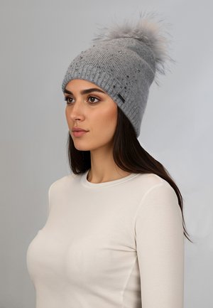 Jeune femme aux longs cheveux noirs portant un bonnet en maille gris clair avec un pompon duveteux et de petites perles décoratives, top à manches longues blanc.