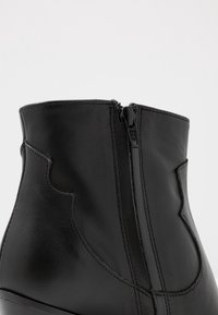 Acercamiento de una bota de cuero negra hasta el tobillo que muestra una cremallera lateral y costuras detalladas en la superficie lisa.