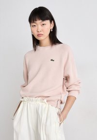 Sweatshirt rosa claro feito de tecido macio, com gola redonda e emblema de logotipo verde. Combinado com calças brancas de cintura elástica.