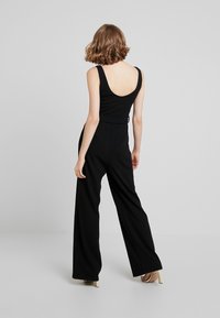Mono negro con cuerpo ajustado y pantalones de pata ancha. Presenta un escote en la espalda y tirantes. Estilizado con tacones metálicos altos.