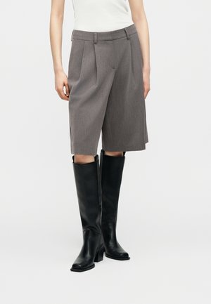 Kvinne iført grå plissert shorts til knelengde kombinert med høye svarte skinnstøvler og en hvit ermeløs topp, stående mot hvit bakgrunn.