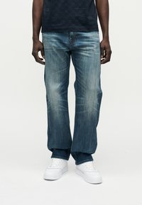 Μπλε τζιν παντελόνι με ευθεία σχεδίαση ποδιού, με ξεβάμματα στους μηρούς και μέση με κουμπί. Φοριέται με λευκά αθλητικά παπούτσια.